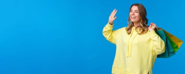Neşeli, mutlu ve rahat genç sarışın kız, elinde alışveriş poşetleri, el sallıyor, arkadaşına gülümsüyor, merhaba diyor, alışveriş merkezinde arkadaşını görüyor, tatil için bir şeyler alıyor, mavi arka plan..