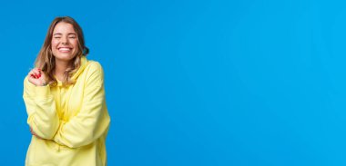 Neşeli kaygısız mutlu kız mutlu ve mutlu, gözleri kapalı mükemmel bir gülümsemeyle gülümseyen, saçlarının teline dokunan, mavi arka planda sarı kapüşonlu bir kız..