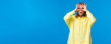 Çok ilginç. Meraklı ve şaşırmış sarışın kız ilginç bir şey görüyor, ağzı açık hayret ediyor, el gözlükleriyle bakıyor ve mavi arka planda dikiliyor..