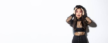 Cadılar Bayramı kostümü giymiş korkmuş ve endişeli Asyalı bir kadın resmi. Korkmuş ya da endişeli görünüyor, el ele tutuşup kameraya bakıyor, beyaz arka planda panikliyor..