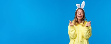 Tatiller, gelenekler ve kutlama konsepti. Neşeli Avrupalı kız, Paskalya bayramını kutluyor, tavşan kulağı takıyor Koreli kalbi işareti yapıyor ve gülümsüyor, mavi arka planda duruyor..