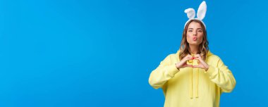 Tatiller, gelenekler ve kutlama konsepti. Sarı kapüşonlu ve tavşan kulaklı komik ve sevimli sarışın kız kalp hareketi yapıyor, mwah öpücük ifadesi yapıyor, sevgi ve pozitiflik yayıyor..