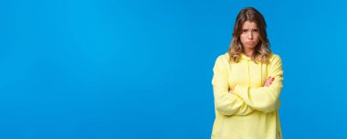 Kasvetli ve huysuz tatlı sarışın genç kız alnının altından bakar ve kameraya somurtur, alınır ya da üzülür, çapraz el ele tutuşur özür bekler, mavi arka plan.