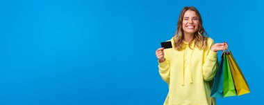Mutlu gülümseyen güzel kız kredi kartını kullanıyor, alışveriş için para yatırıyor, elbiseli çantaları tutuyor ve sırıtıyor, sonunda yaz tatiline hazırlanıyor, mavi arka plan..