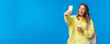 Yaşam tarzı, üniversite ve eğitim konsepti. Neşeli sarışın kız öğrenci ya da gezgin sırt çantalı selfie çekiyor ve kağıt bardakta kahve içiyor, cep telefonu kamerasına göz kırpıyor..
