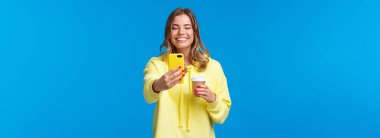 Neşeli, güzel, sarışın, kısa saçlı, sarı kapüşonlu kadın. En sevdiği kafeden özel kahve alıp selfie çekiyor. Elinde cep telefonu kamerasıyla gülümsüyor..
