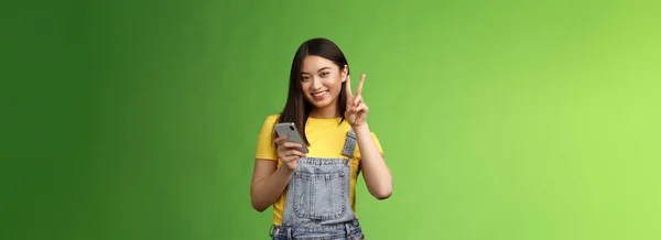 かわいい柔らかいアジアの女性は スマートフォンを保持し 勝利の平和のサインを表示するには 緑の背景の近くにカメラを喜んで 屈託のないポーズを見て オンラインプレゼントに勝つ 喜んでビートスコアゲーム — ストック写真