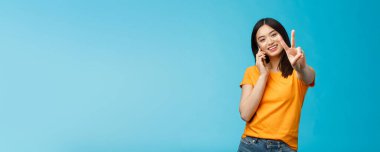 Karizmatik şirin kız olumlu haberleri paylaşıyor akıllı telefon gösterisi barış, zafer işareti mutlu bir şekilde gülümsüyor, mavi arka planda duruyor hoş bir telefon görüşmesi alıyor, telefon aracılığıyla anlaşmaya varıyor..