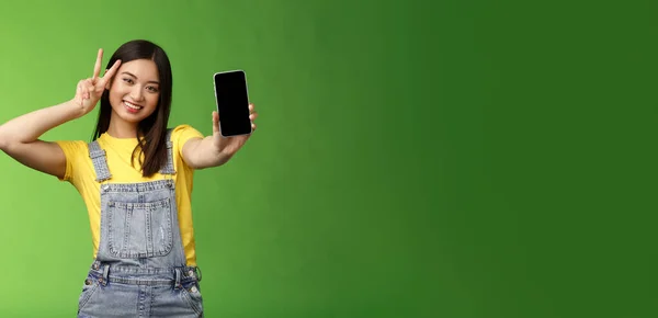 可爱无忧无虑的亚洲黑发在智能手机屏幕上显示应用 让胜利和平的标志 快乐的微笑 吹嘘社交媒体的普及程度 追随者数量 绿色背景 — 图库照片