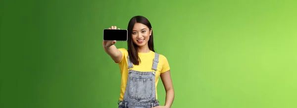 魅力的な陽気なアジアのブルネットの女性は スマートフォンゲームを紹介し 電話を水平方向に保持し 携帯電話のディスプレイを表示し 喜んで笑顔 アプリのダウンロード 緑の背景をお勧めします — ストック写真