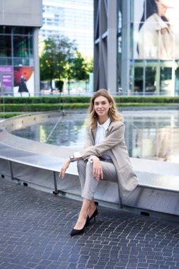 Takım elbiseli başarılı iş kadınının dikey çekimi, çeşme ve iş yerinin yanında oturuyor, kameraya özgüvenle gülümsüyor..