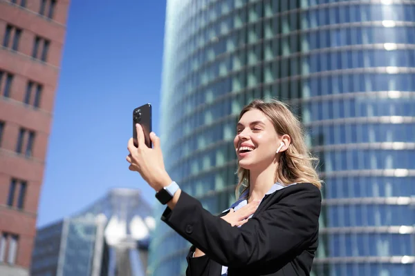 Portrait of smiling corporate woman video call on street, holding mobile phone and waving at smartphone camera, wearing suit.