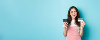 İnternetten alışveriş yapan, plastik kredi kartı gösteren, akıllı telefon tutan, mavi arka planda duran şık genç bir kadın..