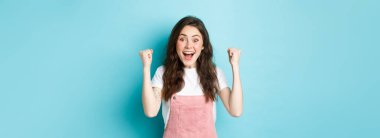 Kazanan o. Neşeli genç bir kadın zaferi kutluyor, hedefe ya da başarıya ulaşıyor, neşeli bir gülümsemeyle evet diye bağırıyor ve zaferi kutlamak için ellerini kaldırıyor, mavi arka plan..