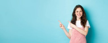 Yazlık kıyafetli mutlu bir genç kızın portresi, sevimli bir allık ve parlak bir makyajı var, fotokopi alanını işaret ediyor, logo reklamını gösteriyor, mavi arka plan..