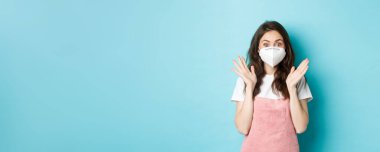 Covid-19, aşı ve karantina konsepti. Tıbbi solunum cihazındaki heyecanlı ve şaşırmış genç kadın, Coronavirus 'un yüz maskesi, el çırp ve kameraya bak, mavi arka plan.
