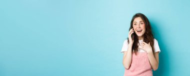 Güzel kız akıllı telefonda konuşuyor ve şaşırtıcı haberler alıyor. Heyecanlı ve şaşırmış görünüyor, telefonla konuşuyor, mavi arka planda mutlu duruyor..