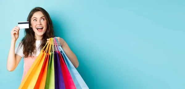 快乐的黑发女孩 带着塑料信用卡和购物袋的购物狂 站在蓝色的背景上 看着商店的标志或横幅 看起来像在做梦 — 图库照片