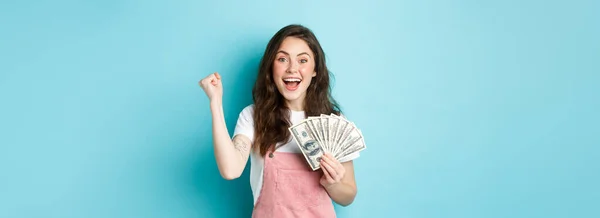 Excited smiling girl fist pump and hold money prize, winning cash, receive income from something, standing happy against blue background.