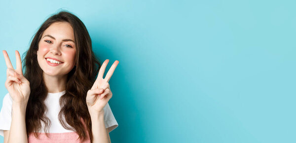 Close up portrait of attractive positive girl saying cheeze, smiling and showing v-signs peace, standing over blue background.