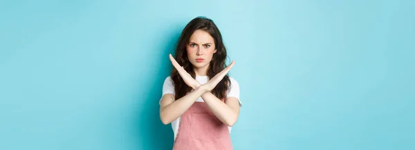 Image of young disappointed woman frowning, making cross sign and sulking, showing stop gesture, say no, forbid bad action, standing over blue background.
