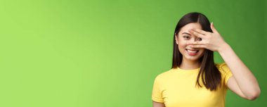 Neşeli, hevesli, tatlı Asyalı esmer kız geniş gülümseyen, dişlek, sırıtan, parmaklarının arasından bakan, büyüleyici ifade, yeşil arka plan. Boşluğu kopyala