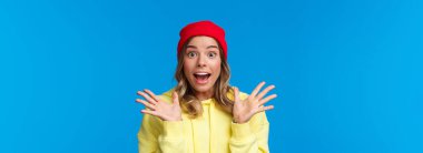 Heyecanlı, güzel Avrupalı kadın harika haberleri açıklıyor, şaşırmış, el sıkışan, büyülenmiş ve kameraya bakan arkadaşlarıyla konuşuyor, kırmızı bereli mavi arka planda duruyor..