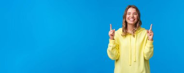 Sarı kapüşonlu, mutlu, mutlu, mutlu, sarı saçlı kız, parmaklarını havaya kaldırarak gülüyor, kaygısız, ürün öneriyor, abonelik tanıtımı yapıyor veya mavi arka planda şirket afişi asıyor..