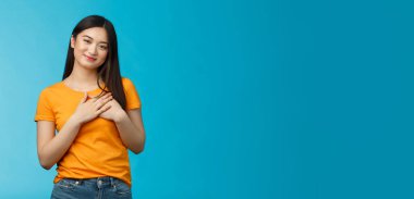 Sevecen, dost canlısı görünümlü kısa saçlı Asyalı kız göğüs avuçlarına dokun, hayran ve şefkatle bak, neşeyle gülümse, iç açıcı sahne, mavi arka plan.