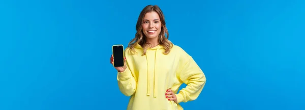 Waist-up portrait good-looking blond girl in yellow hoodie promote smartphone application, show mobile phone display and smiling camera as recommend subscribe or download app.