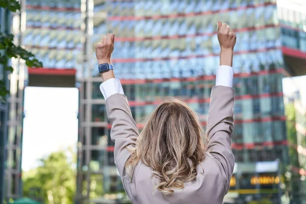 Corporate people. Rear view of businesswoman raising hands up in celebration, triumphing, achieve goal and rejoicing, standing on street.