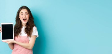 Eğlenceli güzel kız boş dijital tablet ekranı gösteriyor ve kameraya heyecanla gülüyor, harika tanıtım teklifleri sunuyor, mavi arka planda duruyor..