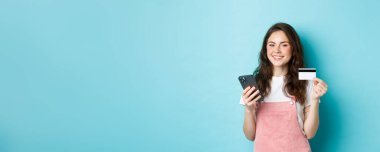 Çevrimiçi ödeme yapan genç bir kadın, elinde kredi kartı ve akıllı telefonla online alışveriş yapıyor, kameraya tatmin olmuş görünüyor, mavi arka planda duruyor..