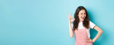 Neşeli cazibeli bir kadının göz kırpması ve iyi bir şeyi tavsiye etmek ve onaylamak için tamam işareti göstermesi, evet demek, iyi seçimi övmek, mavi arka plan..
