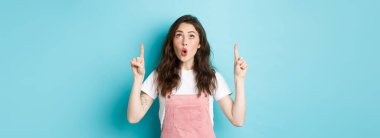 Vay canına, çok havalı. Siyah kıvırcık saç stili ve cazibeli etkileyici genç kız, şaşkın ve heyecanlı görün, reklamlara işaret et ve dik dik bak, mavi arka plan.