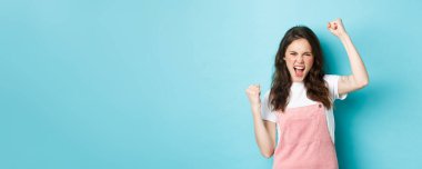 Neşeli genç bir kadın takımı destekliyor, maç seyrediyor ve tezahürat yapıyor, yumruk tokuşturarak el kaldırıyor, insanları cesaretlendirmek için bağırıyor, mavi arka planda duruyor..