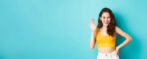 Summer holidays and emotions concept. Cheerful friendly woman with perfect body, waving hand and saying hello, making hi gesture, greeting you, standing over blue background.