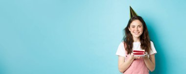 Tatiller ve kutlamalar. Parti şapkalı neşeli doğum günü kızı elinde pasta, gülümsüyor, mum üzerinde dilek diliyor, mavi arka planda duruyor..