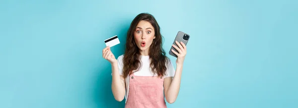 惊讶的年轻女子说 兴奋地盯着相机 拿着手机和信用卡 站在蓝色的背景上 — 图库照片