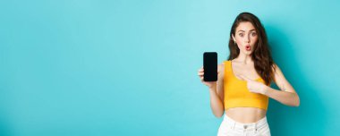 Akıllı telefon uygulaması hakkında soru soran, mobil ekrana bakan ve mavi arkaplana karşı duran meraklı kız. Boşluğu kopyala