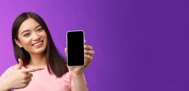 Yakın plan, neşeli, karizmatik Asyalı kadın başını eğiyor. Güzel, mevcut uygulama, akıllı telefon ekranını göster. Uygulamayı tanıt, işaret eden telefon, işaret parmağı ekranı, mor arka plan..