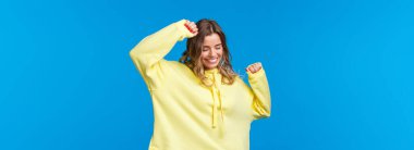 Mutlu ve rahat, kaygısız, beyaz sarışın kız partide eğleniyor, müzik dinliyor, el kaldırıyor ve neşeyle dans ediyor, kapalı gözlerle gülümsüyor, mavi arka planda duruyor..