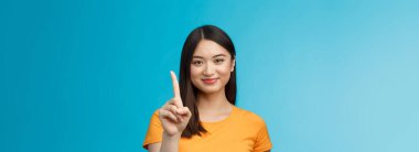 Yakın plan, çekici, kısa saçlı, çekici Asyalı kadın. Bir numara, işaret parmağını uzat, neşeyle gülümse, dostça gülümse. Mavi arka planda dur..