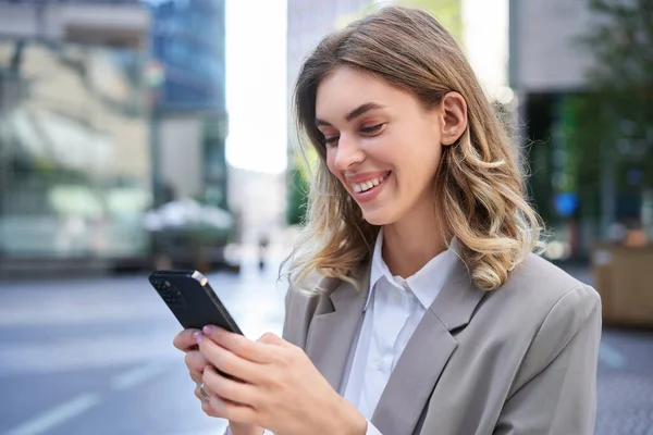 Portrait of smiling businesswoman chat in app, looking at smartphone with pleased face, order delivery to office, reading message on mobile phone.
