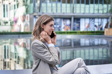Genç başarılı iş kadını ofis binasının dışında oturuyor ve cep telefonuyla konuşuyor, takım elbise giyiyor ve topuklu ayakkabı giyiyor..