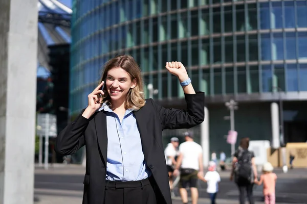 Excited saleswoman receive good news on phone and rejoicing, walking on street and celebrating, making fist pump while on telephone.