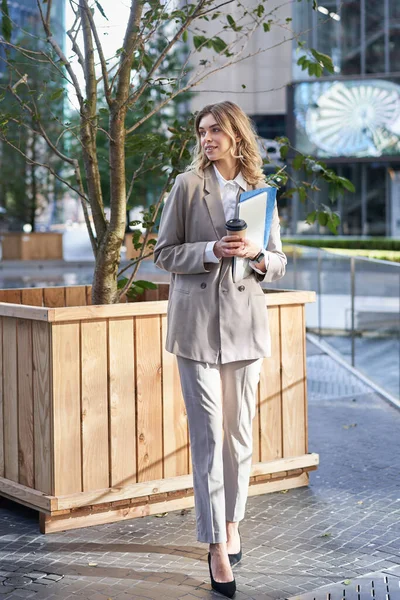 Stylish corporate woman in suit, standing on street with documents, work laptop and coffee, waiting near business center.