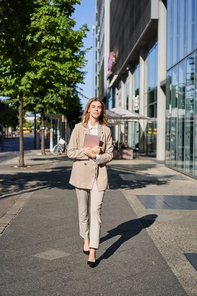 Vertical shot of businesswoman walking on street with digital tablet, going to work, wearing beige suit and high heels.
