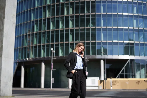 Young corporate woman in suit walking on street and talking on mobile phone. Businesswoman having telephone conversation while going to work.