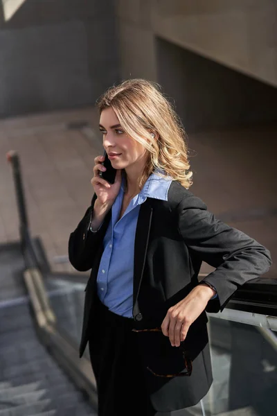 Portrait of corporate woman standing on escalator, making a phone call, using smartphone, walking in city.